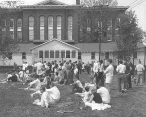 A group of students laying down on a grassy field behind a three-story large brick and wood building