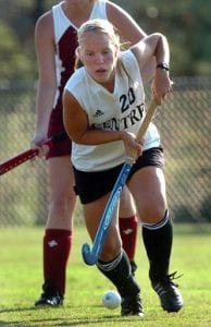 Portrait of a female field hockey athlete on a grass field with a field hockey stick in hand moving a ball.